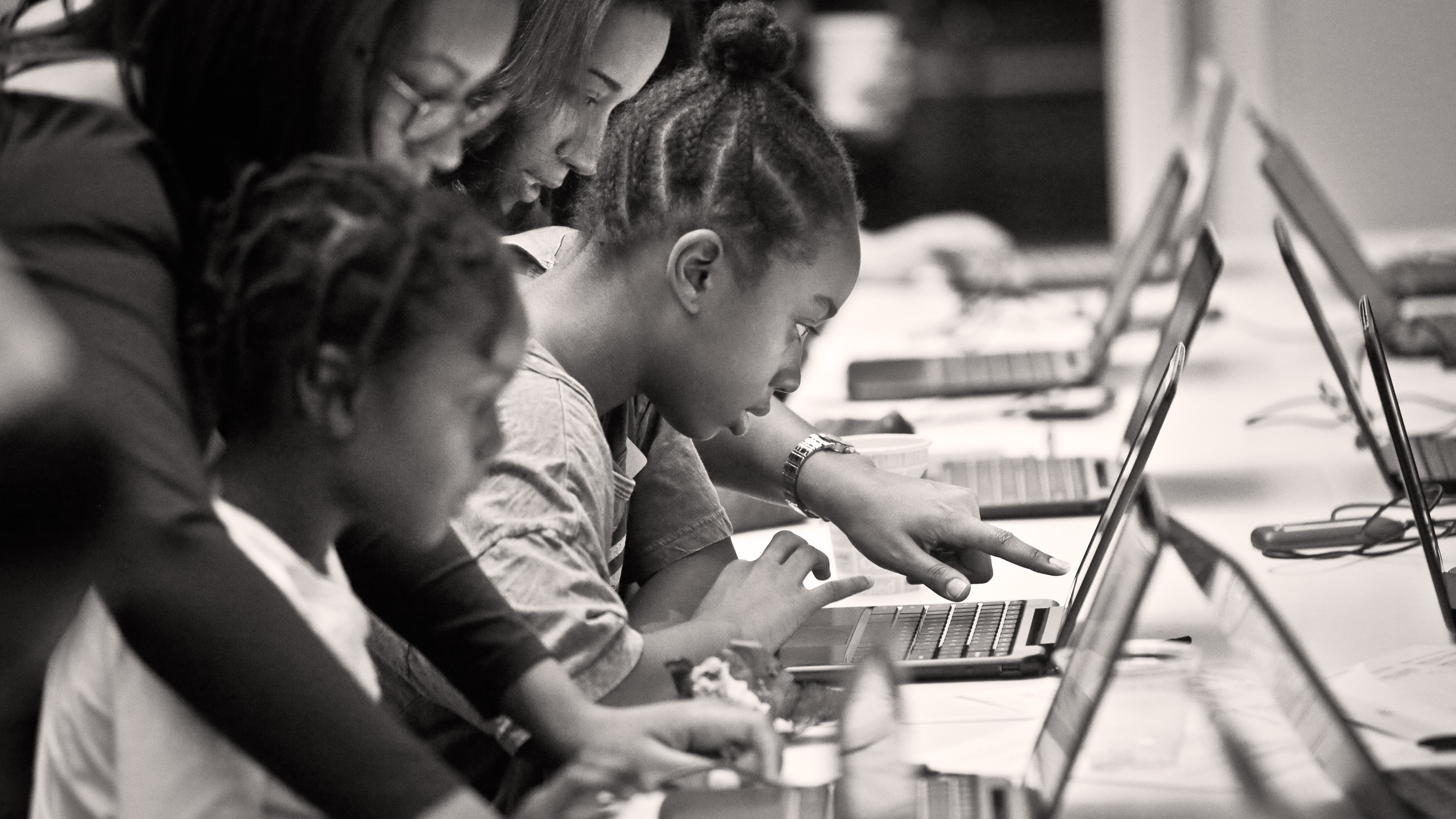 Women lean over to help young girls working together on their laptops at a long table